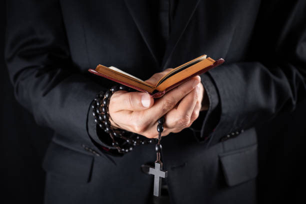 as mãos de um padre cristão vestiram-se na terra arrendada preta um crucifixo e lendo o livro do testamento novo. - religious text cross protestantism reading - fotografias e filmes do acervo