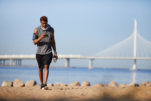 Serious handsome young black guy with headphones on neck texting sms while walking over beach in summer