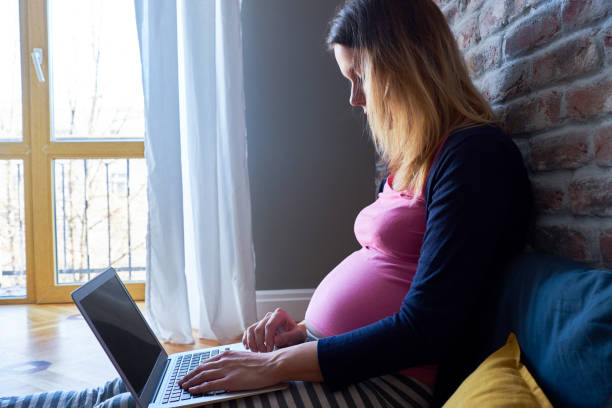Pregnant woman working at the computer, sitting on the floor in her pajamas in daylight stock photo