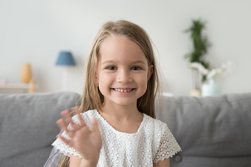 Smiling little girl looking at camera, waving hand, greeting, making video call to relatives, recording vlog for channel, head shot portrait of cute child, sitting on cozy sofa at home