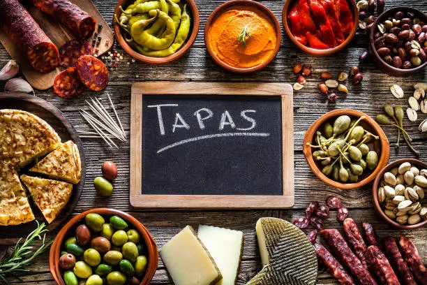 Spanish food frame: tapas. Chorizo, manchego cheese, spanish omelette, olives, capers, jalapeño peppers, pimientos piquillo, pistachios, peanuts and sun dried tomatoes shot from above on dark rustic wooden table. The composition is arranged all around the border of an horizontal frame and at the center is a blank blackboard with the text "Tapas". Predominant colors are red and brown. Low key DSRL studio photo taken with Canon EOS 5D Mk II and Canon EF 100mm f/2.8L Macro IS USM.