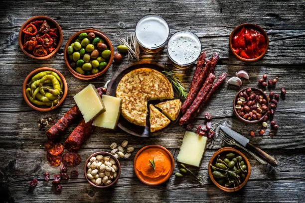 Spanish food: tapas and beer. Chorizo, manchego cheese, spanish omelette, olives, capers, jalapeño peppers, pimientos piquillo, pistachios, peanuts and sun dried tomatoes with two beer glasses shot from above on dark rustic wooden table. The food is in clay bowls. Predominant colors are red and brown. Low key DSRL studio photo taken with Canon EOS 5D Mk II and Canon EF 70-200mm f/2.8L IS II USM Telephoto Zoom Lens