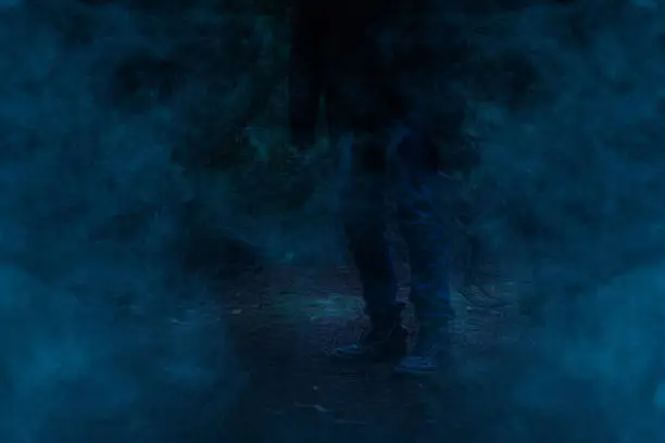 Photo of a mysterious man is standing in a night forest covered with blue ghostly fog