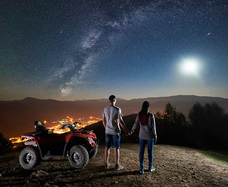 Back view of young couple tourists with atv quad motorbike on the top of mountain. Man and woman holding hands, enjoying night sky full of stars, full moon, Milky way, luminous city on background