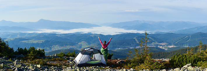 Panoramic view of camping on the top of mountain on summer morning. Back view of young hiker woman having a rest beside tourist tent, holding hands lifting up. On beautiful foggy mountains background.