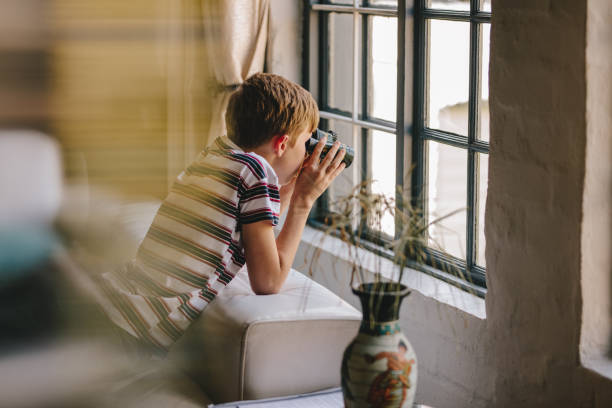 любопытный мальчик смотрит в окно с биноклем - curiosity searching discovery home interior стоковые фото и изображения