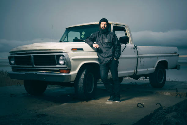 A serious looking bearded man, standing with a 50 year old pickup truck at the shoreline at dusk.