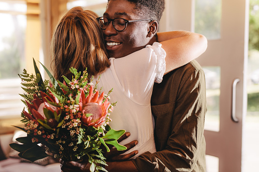 Young african man hugging his girlfriend standing in cafe. Young guy expressing his love for his lady giving flowers and a warm hug during a coffee shop date.