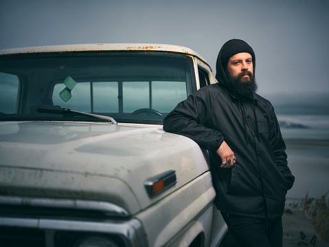 A serious looking bearded man, standing with a 50 year old pickup truck at the shoreline at dusk.