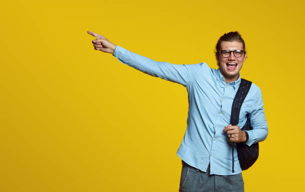 istock Smiling young man with eyeglasses, holding backpack and pointing finger away isolated on a yellow background. 1133645511