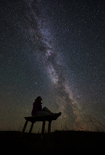 Dark silhouette of female hiker resting at summer night camping in the mountains. Young woman sitting on a bench, enjoying view of night sky full of stars and Milky way. Astrophotography