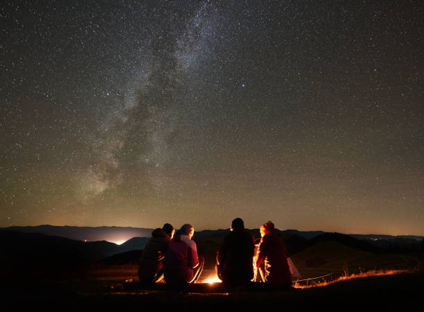 Friends resting beside camp, campfire under night starry sky Night summer camping in the mountains. Back view group of four friends tourists sitting on a bench made of logs together around campfire under amazing night starry sky full of stars and Milky way. campfire stock pictures, royalty-free photos & images