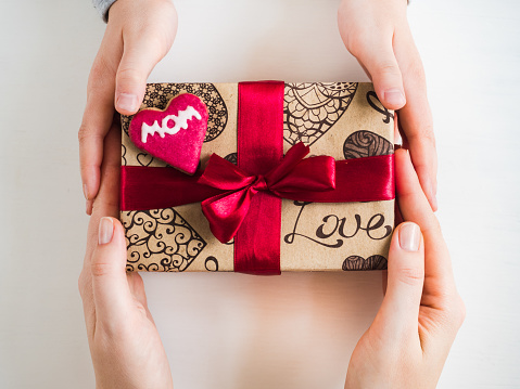 Hands of a younger daughter, beautiful box with a gift, red ribbon and glazed cookies with the word MOM on a white, isolated background. Top view, close-up. Preparing for the holidays