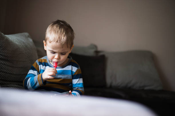 Growing up in a technology-based world Two years old boy is eating a Lollipop while using mobile phone at home. Candy bad for kid teeth. image based social media photos stock pictures, royalty-free photos & images