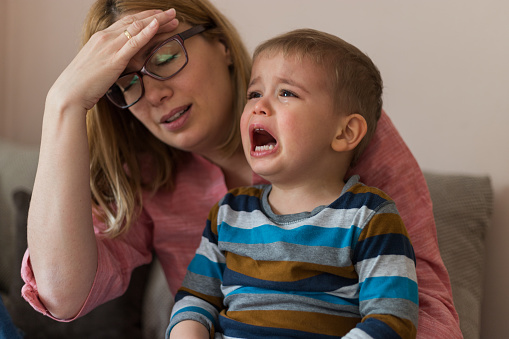 Tired, desperate mother is holding her two year old son, crying.Negative human emotion face. Upset toddler boy. Problem child. Depression, stress or frustration.