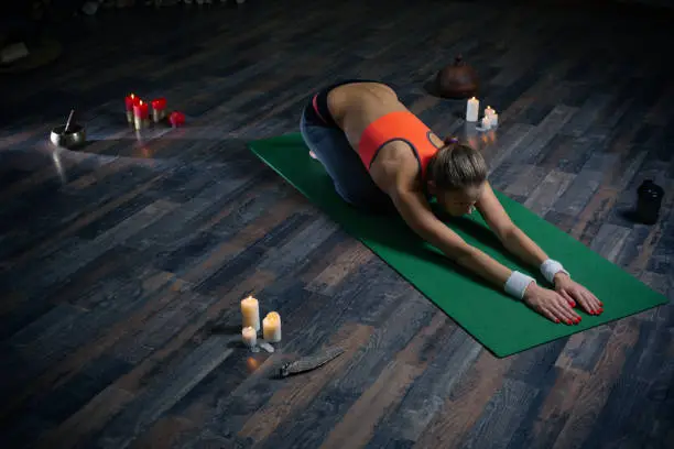Full length of yoga training and young woman bending to the ground while practicing asana on the yoga mat with candles around her