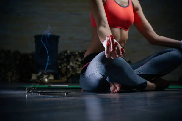 Slim lady sitting on the yoga mat and putting hands on the crossed legs while meditating with incense stick on the floor