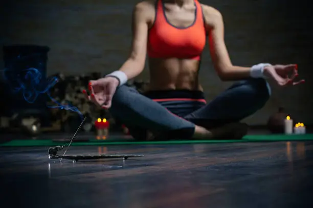 Relaxed young woman meditating on the yoga mat with hands on knees and having incense stick on the floor in front of her