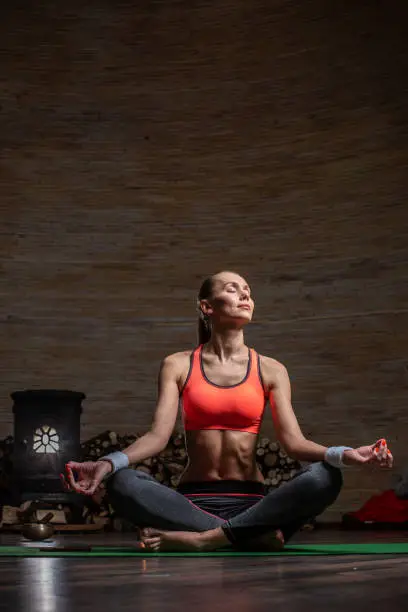 Beautiful young woman in sporty clothes sitting on the green yoga mat with crossed legs and closing her eyes while meditating. Firewood on the background