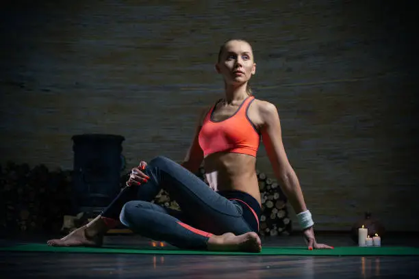 Beautiful slim young lady in sporty clothes sitting on the yoga mat and looking into the distance while twisting her legs during the training
