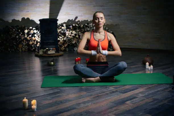 Beautiful calm lady sitting in dark room with fireplace and firewood on the background while meditating with closed eyes