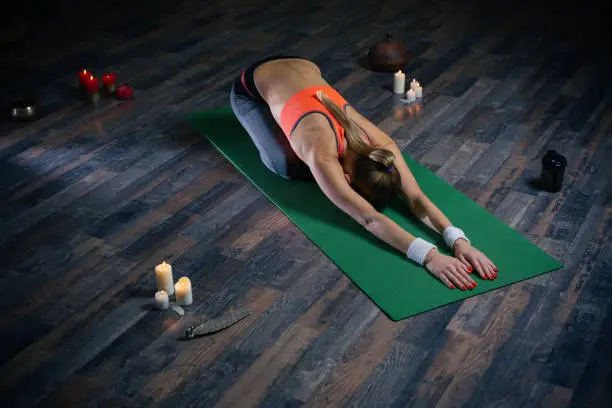 Young slim lady wearing sporty clothes and bending her back while training on the yoga mat with candles around her