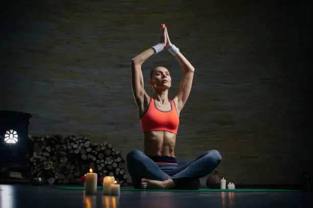 Beautiful slim lady sitting with legs crossed and putting hands above the head while having firewood behind the back and candles on the floor