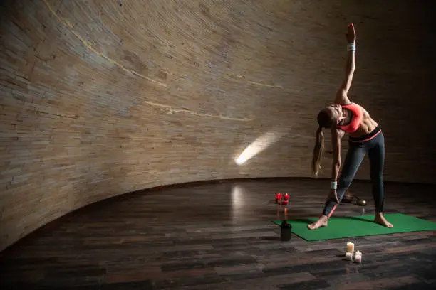 Sporty fit lady standing on the yoga mat with one hand up and practicing the triangle pose in spherical room with candles on the floor