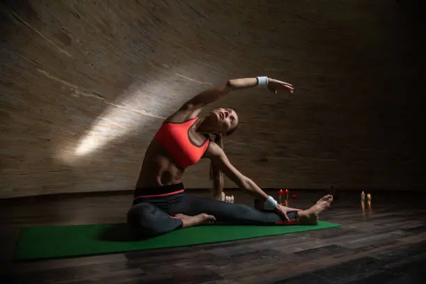 Peaceful young beautiful woman sitting on the yoga mat and putting one hand over the head while doing yoga asana with candles on the background