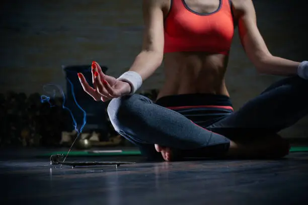 Close up of incense stick on the floor in front of the young fit meditating woman