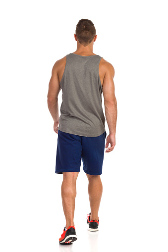 Rear view of walking fit man in gray tank top, blue shorts and orange sneakers. Full length studio shot isolated on white.