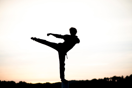 Anonymous silhouette of a young man kicking and punching as he practices martial arts at sunset.