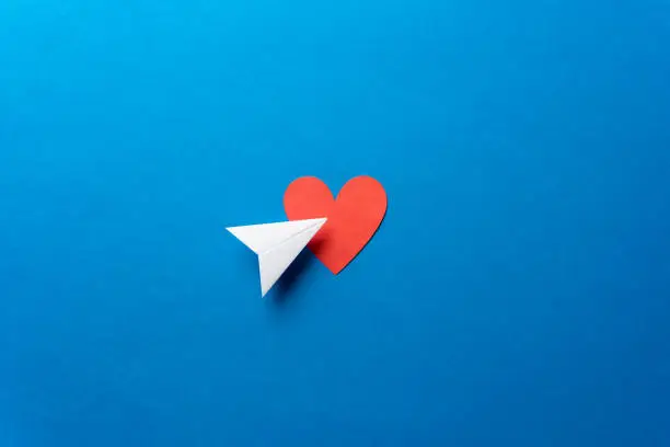 Photo of Paper airplane with red heart shape on blue background. Sharing and send symbol concept. Airplane flight transport sign. Landing page concept.