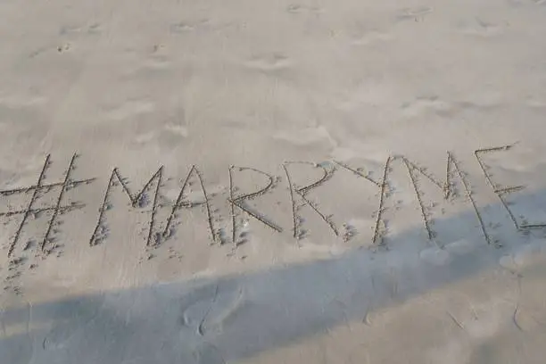 Image of hashtag tag writing on sunny beach in the soft sand, light by the early morning sunshine. Written using a stick by hand, with Marryme words as capital letters by the gentle lapping waves of the sea on the seashore. Picture taken at Palolem Beach, Goa, India, during the early morning golden hour without tourists on holiday vacation / beachgoers.  Concept photo of Marryme handwriting in golden sand with modern hashtag prefix