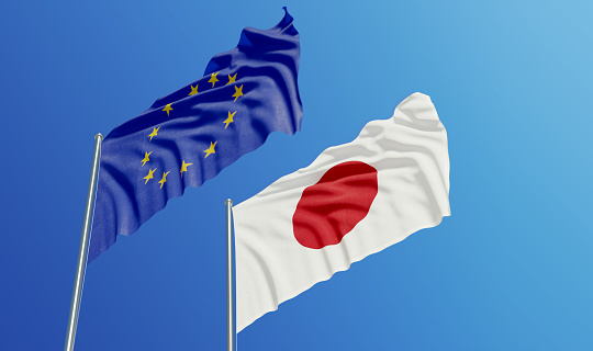 European Union and Japanese flags are waving with wind over blue sky. Low angle view. Dispute and conflict concept. Horizontal composition with copy space.