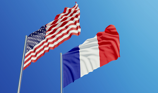 American and French flags are waving with wind over  blue sky. Low angle view. Dispute and conflict concept. Horizontal composition with copy space.
