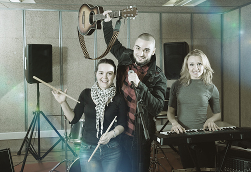 Three happy cheerful positive smiling bandmates posing together with musical instruments in rehearsal room