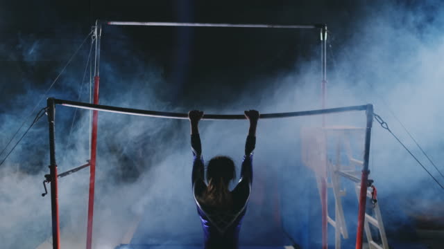 Competitions in gymnastics. A woman walks to the crossbar and performs dips flip-flops in slow motion on a dark background in backlight. Slow motion