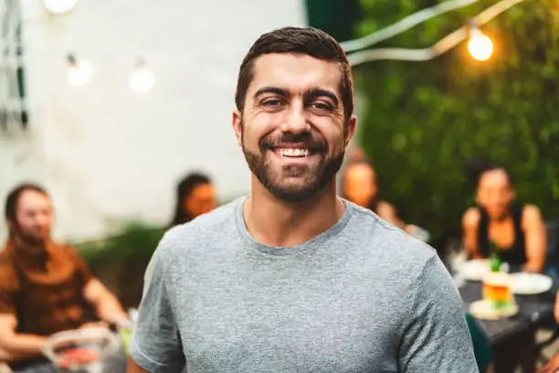 Portrait of cheerful bearded man in yard. Happy male is enjoying garden party with friends in background. He is wearing casuals.