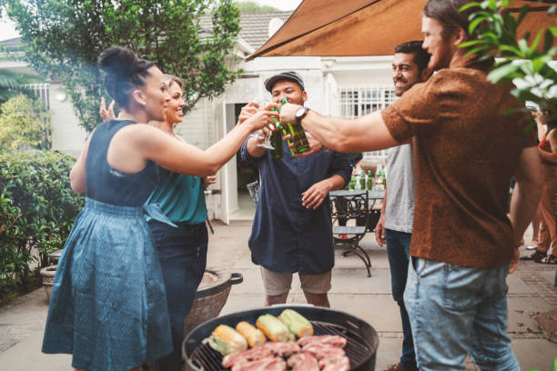 Happy friends toasting drinks by barbecue in yard Happy friends toasting drinks by barbecue. Men and women are enjoying garden party at backyard. They are wearing casuals. south african braai stock pictures, royalty-free photos & images