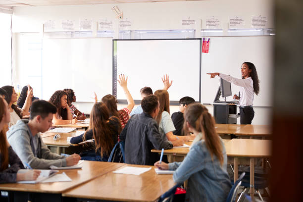 Female High School Teacher Asking Question Standing By Interactive Whiteboard Teaching Lesson Female High School Teacher Asking Question Standing By Interactive Whiteboard Teaching Lesson hand raised classroom student high school student stock pictures, royalty-free photos & images