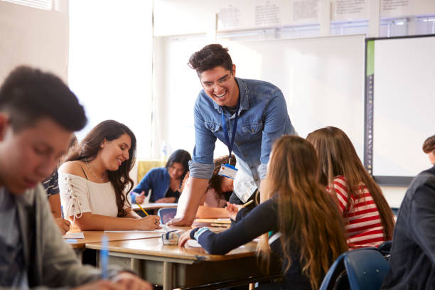 Male High School Teacher Standing By Student Table Teaching Lesson Male High School Teacher Standing By Student Table Teaching Lesson teacher classroom child education stock pictures, royalty-free photos & images