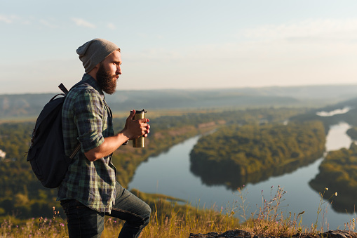 Side view of handsome bearded guy with backpack holding warm thermos and looking away while standing in magnificent countryside