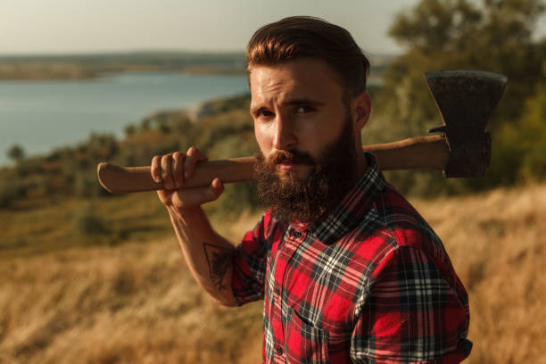 Bearded male with axe in countryside Handsome bearded lumberjack in checkered shirt holding axe on shoulder and looking at camera while standing in amazing countryside lumberjack stock pictures, royalty-free photos & images