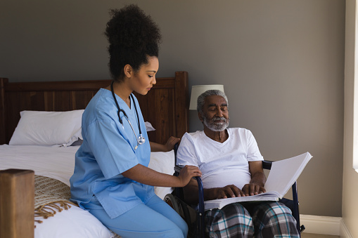 Front view of a blind senior African American man reading a braille with young African American female doctor at home