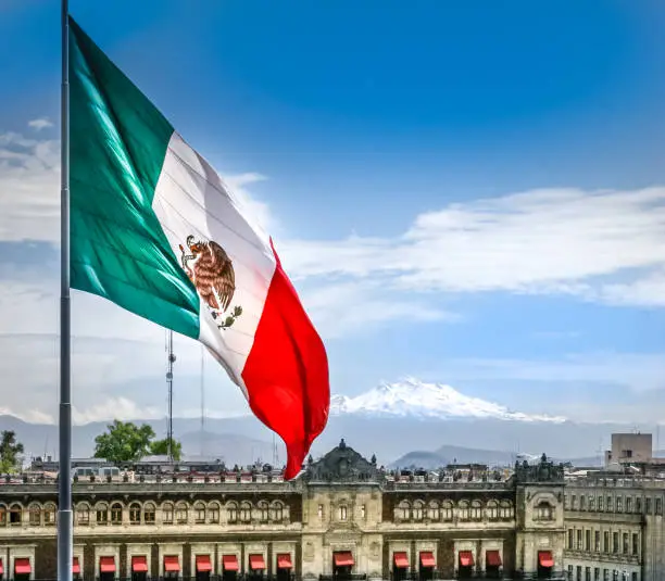 Mexico City, Mexico, Apr 20 - The monumental and permanent flag of Mexico hoisted in the Zocalo of Mexico City, the most great square in the country. At  the bottom the National Palace (Presidential House) and the magnificent Iztaccíhuatl volcano. The Zocalo is the heart of the historic center of Mexico City, declared a World Heritage Site in 1987 by Unesco.