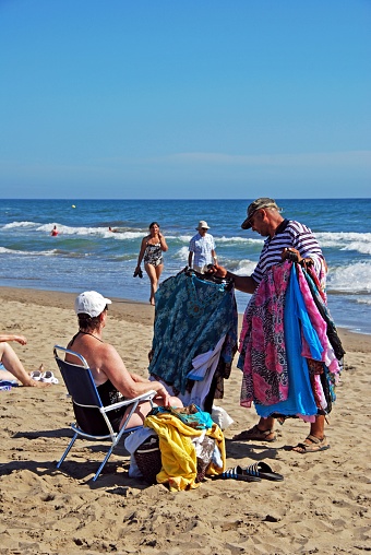 Beach trader showing his goods to a tourist on Playa de la Vibora beach, Elviria, Marbella, Malaga Province, Andalusia, Spain, Western Europe.