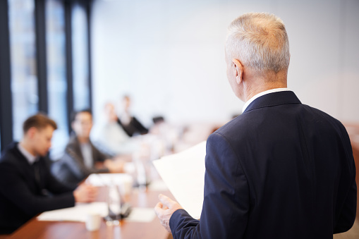 Back view portrait of senior businessman giving speech in conference room talking to employees, copy space