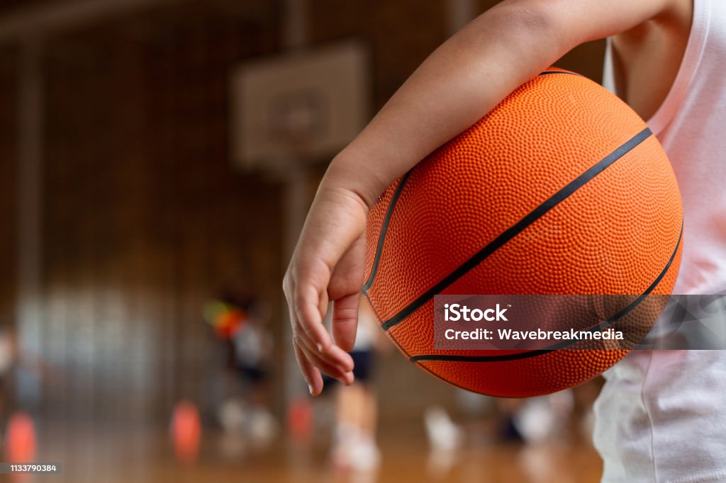 Schoolboy with basketball standing in basketball court Mid section of a mixed-race schoolboy with a basketball under his arm standing in basketball court at school Child Stock Photo