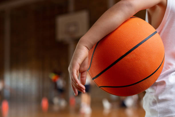 Schoolboy with basketball standing in basketball court Mid section of a mixed-race schoolboy with a basketball under his arm standing in basketball court at school youth basketball indoor stock pictures, royalty-free photos & images